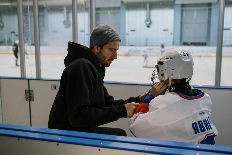A father helps his son with hockey gear at an indoor ice rink.