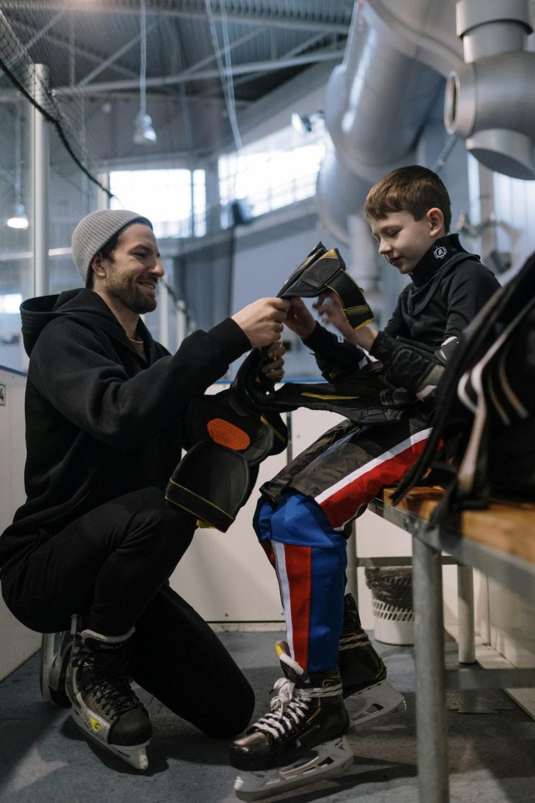 Father assisting son with ice hockey gear in an indoor rink, creating a warm teaching moment.